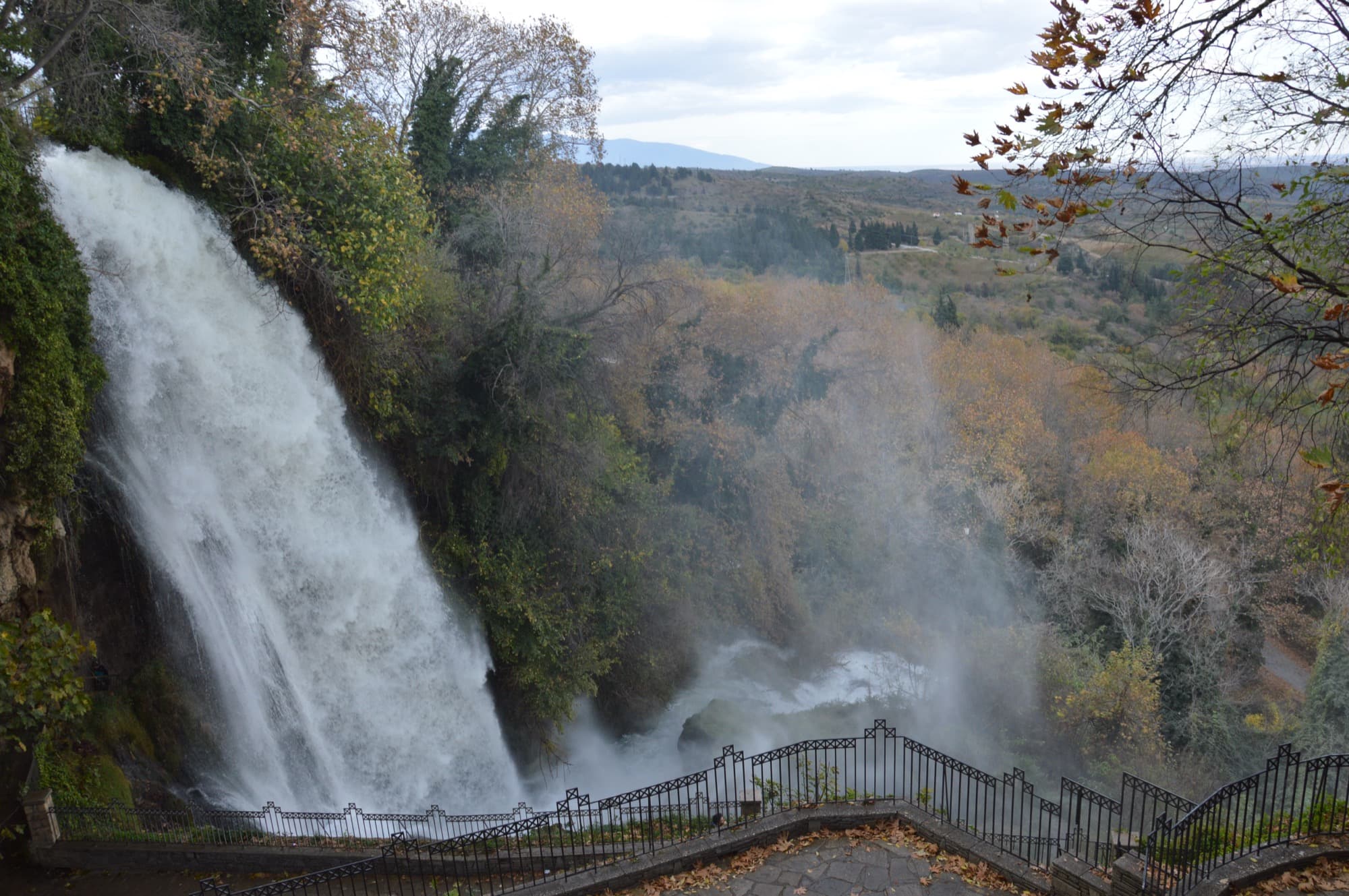 The Karanos waterfall at Edessa drops off the cliff beside a railed stone terrace, autumn trees and the plain beyond.