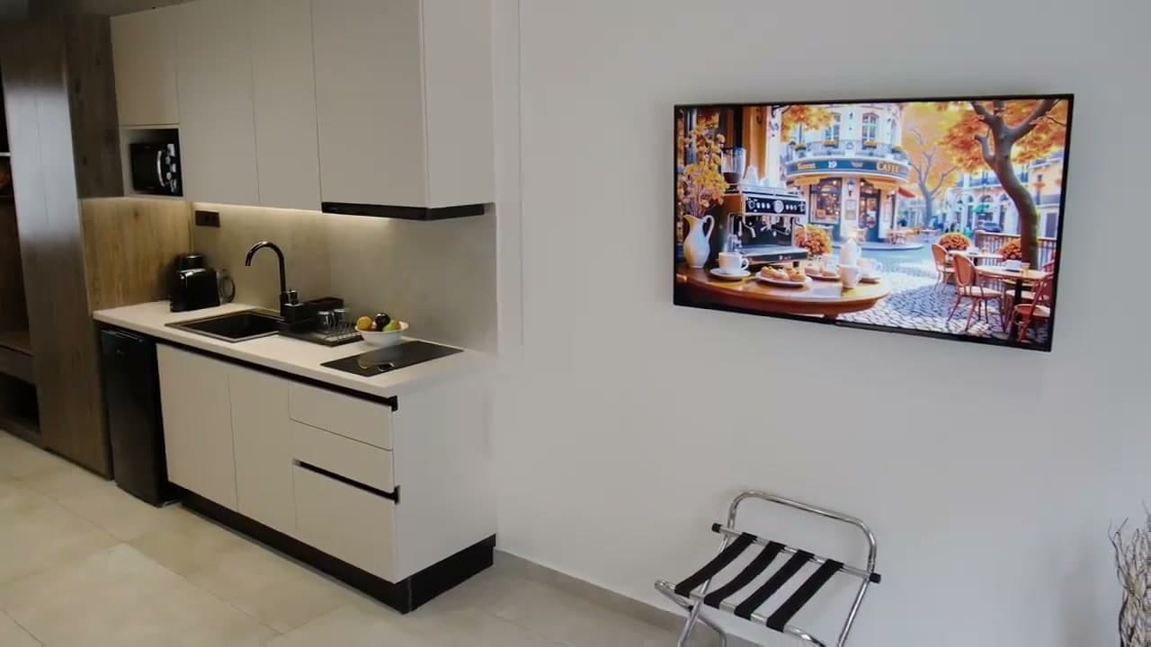 The suite's kitchenette along one wall: white cabinets, a sink with a black tap, an induction hob, a microwave and an under-counter fridge, with a wall-mounted TV opposite